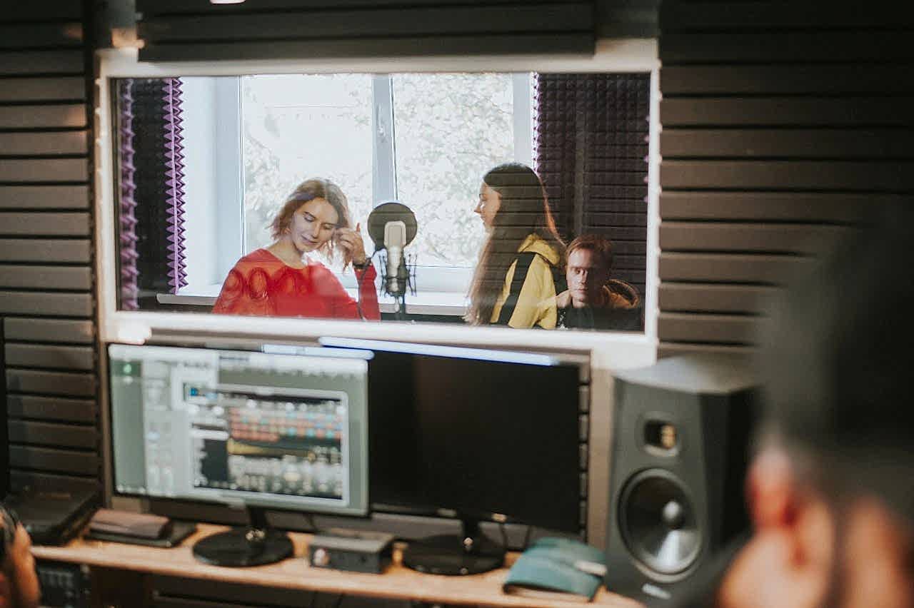 hero-img-01 Artists recording music in a professional studio setting, captured through a glass window.