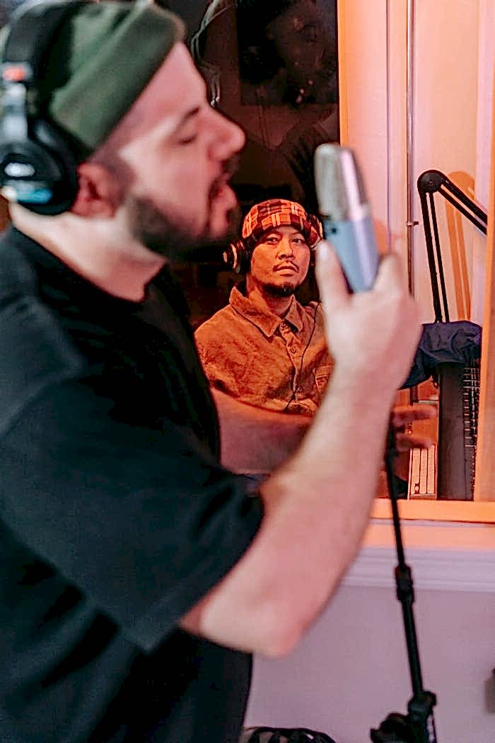services-03 Two men collaborate in a recording studio. One sings into a microphone while the other observes.